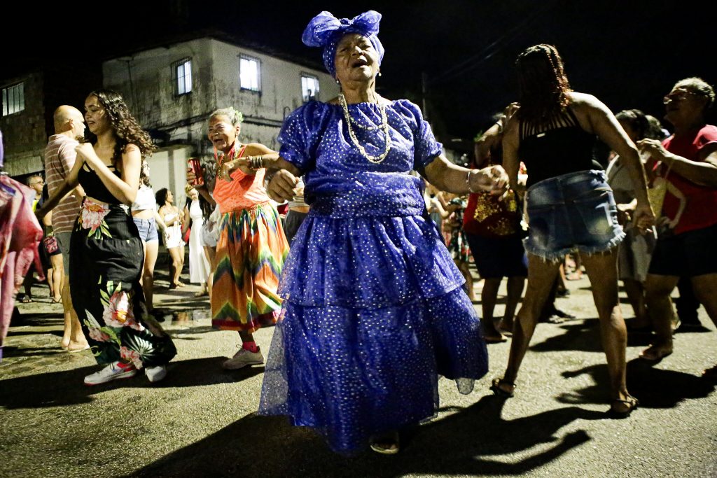Foto colorida de ensaio de maracatu à noite, com passistas em torno de uma mulher negra idosa, vestida com longo vestido azul, de saia armada, dançando no meio da rua.