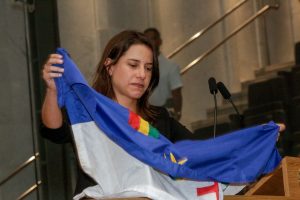 Foto colorida da governadora Raquel Lyra, com semblante sério, abrindo bandeira de Pernambuco na tribuna da Assembleia Legislativa.