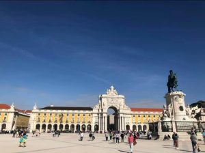 Foto colorida da Baixa de Lisboa, com largo amplo em primeiro plano e construção antiga pintada de amarelo com detalhes brancos sob céu azul sem nuvens. Ao fundo se vê o arco de entrada da rua Augusta ao centro, tendo uma estátua equestre à direita da imagem.