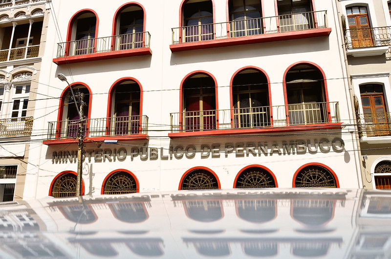 Foto colorida de um um edifício branco com detalhes em vermelho e a inscrição “MINISTÉRIO PÚBLICO DE PERNAMBUCO” na frente. O edifício é refletido no teto de um carro que está estacionado em frente a ele.