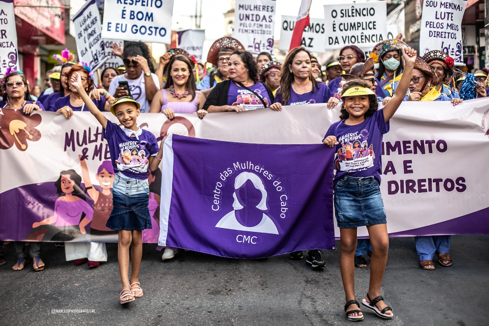 Grupo de mulheres reunidas em caminhada, carregando faixas e cartazes na cor lilás. A frente, duas meninas vestidas com saia, blusa roxa estampada e viseiras, carregando uma faixa com a logo do Centro das Mulheres do Cabo.