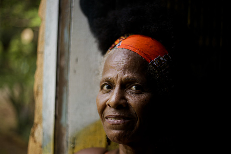 Foto do rosto de Luiza Cavalcante, mulher negra e idosa, usando uma bandana laranja na cabeça. Ela foi fotografada na penumbra, junto a uma janela iluminada.