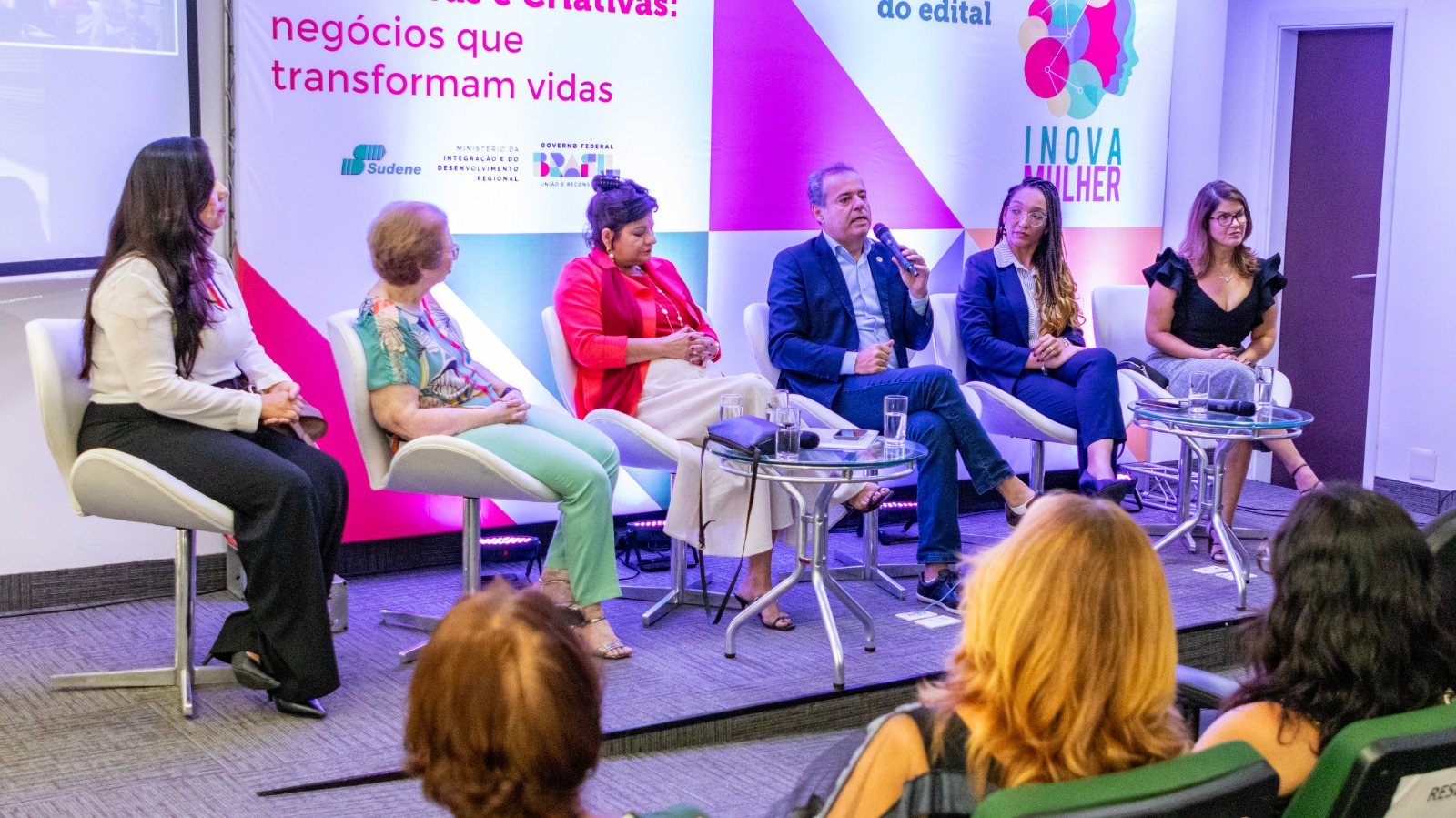 Ladeado por mulheres em tablado que serve como palco tendo um fundo nas cores rosa e branco, Danilo Cabral fala ao microfone.