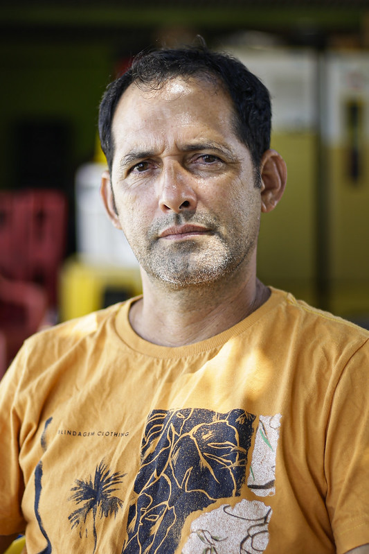 Foto de Jamerson Pereira, homem branco, jovem, de cabelos e olhos escuros, barba mal feita, usando uma camisa laranja com estampas pretas e brancas. O fundo está desfocado, mas a foto foi feita ao ar livre.