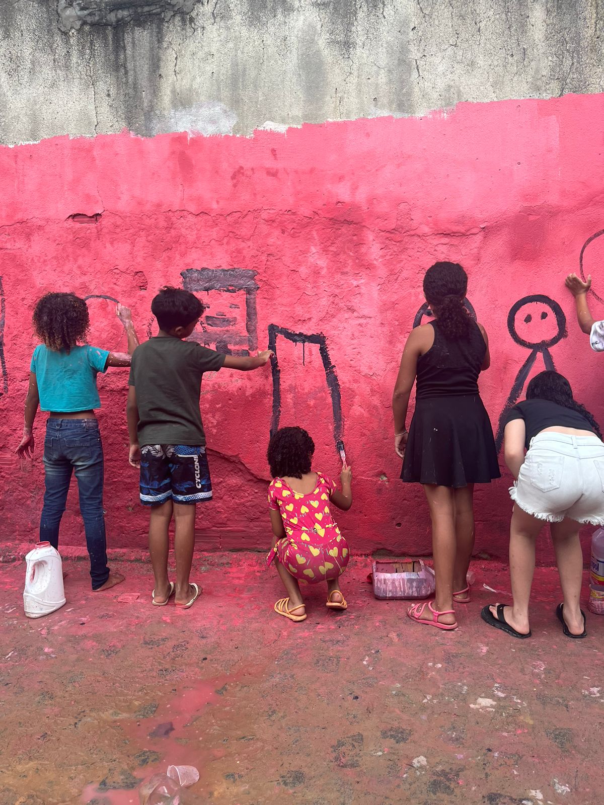 A imagem mostra várias crianças desenhando em uma parede externa. A parede é texturizada e pintada de rosa, com partes desgastadas mostrando a cor cinza do concreto por baixo. As crianças estão focadas em seus desenhos, que incluem formas geométricas e um boneco de palito. É uma cena colorida e animada, com as crianças expressando sua criatividade e se divertindo ao ar livre. A textura da parede e as poças d’água no chão sugerem choveu pouco antes.