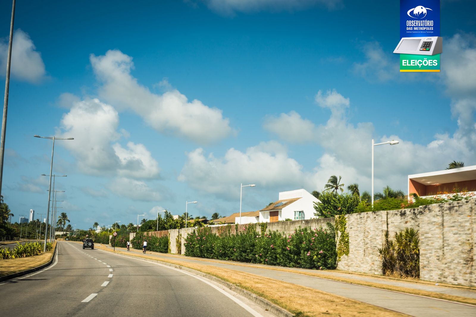 Nesta foto, vê-se uma estrada asfaltada com veículos, cercada por vegetação e casas com muros altos, sob um céu azul com nuvens; no canto superior direito, há um logo e texto do Observatório das Metrópoles.