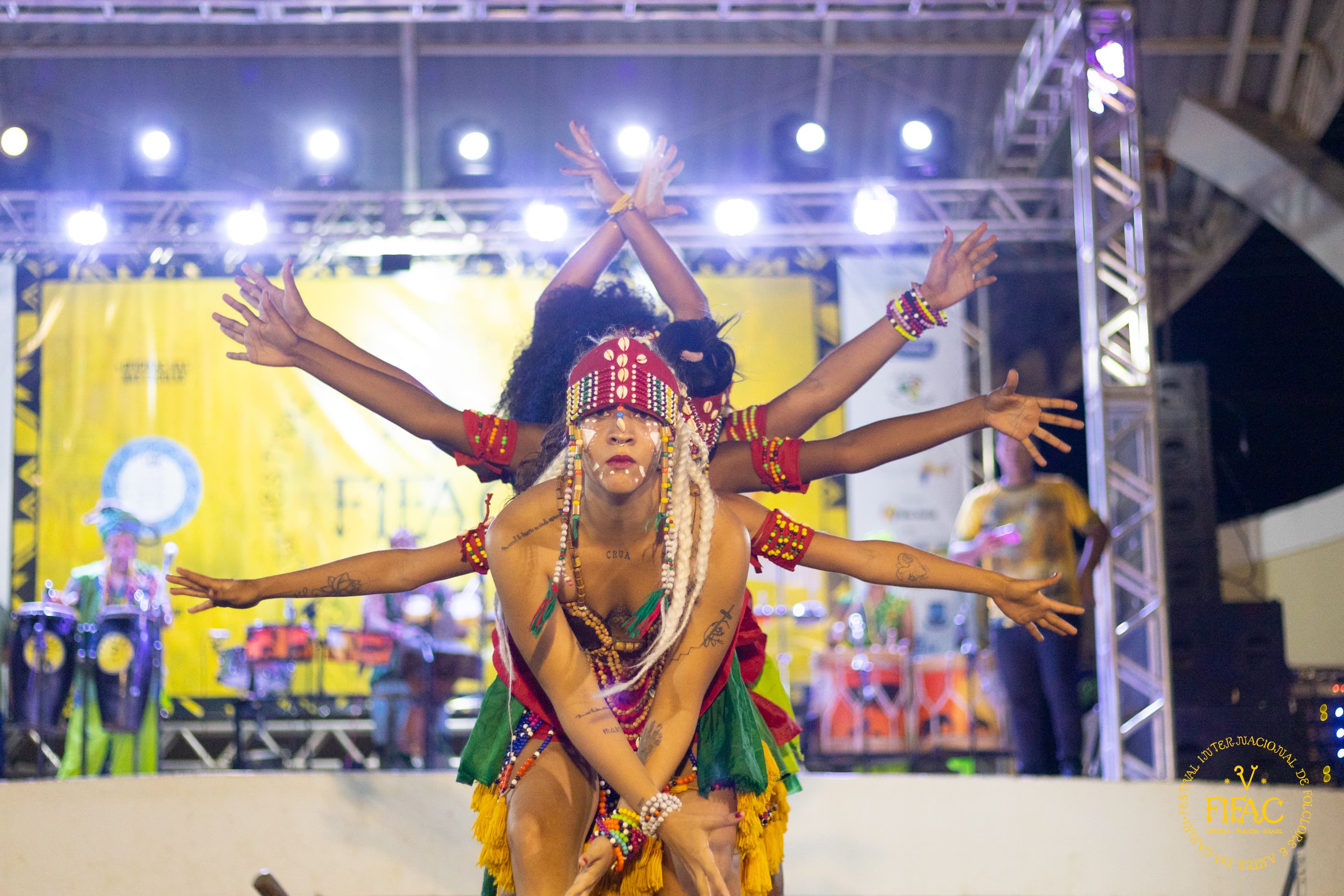 Foto de apresentação noturna do grupo Bacnaré, com jovem negro à frente de um grupo de bailarinos, que usam braços e mãos para simular um efeito de que a pessoa em primeiro plano parece ter vários braços. Ao fundo, se vê um palco iluminado.