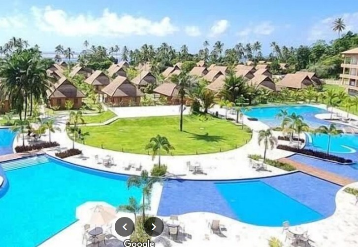 Esta imagem retrata um resort luxuoso com uma atmosfera tropical. Possui várias piscinas interligadas, gramados verdes e bangalôs com telhados que aparentam ser de palha. O céu está claro, sugerindo um dia ensolarado