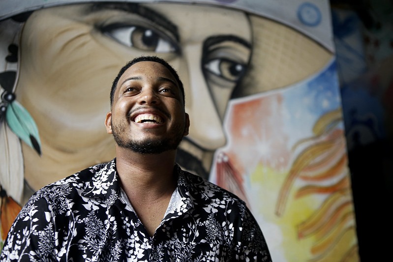 Foto de Nailson Vieira, jovem negro com sorriso largo, olhando para o alto, usando uma camisa estampada com motivos florais em preto e branco. Ao fundo, a parede está pintada com uma imagem de uma pessoa negra, usando chapéu com fundo colorido.