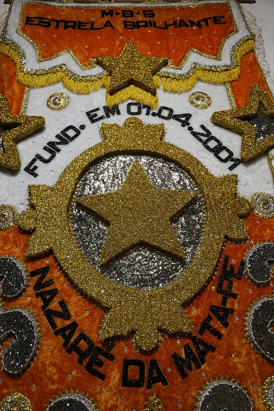 Foto do estandarte do maracatu Estrela Brilhate, com um fundo laranja texturizado. No centro, há uma grande estrela dourada brilhante, cercada por formas que se assemelham a raios de sol. O texto “M.B.S ESTRELA BRILHANTE” está escrito na parte superior do emblema. Abaixo da estrela central, o texto “FUND. EM 01.04.2001 NAZARÉ DA MATAPE” é visível. Pequenas decorações circulares adornam a parte inferior do emblema.