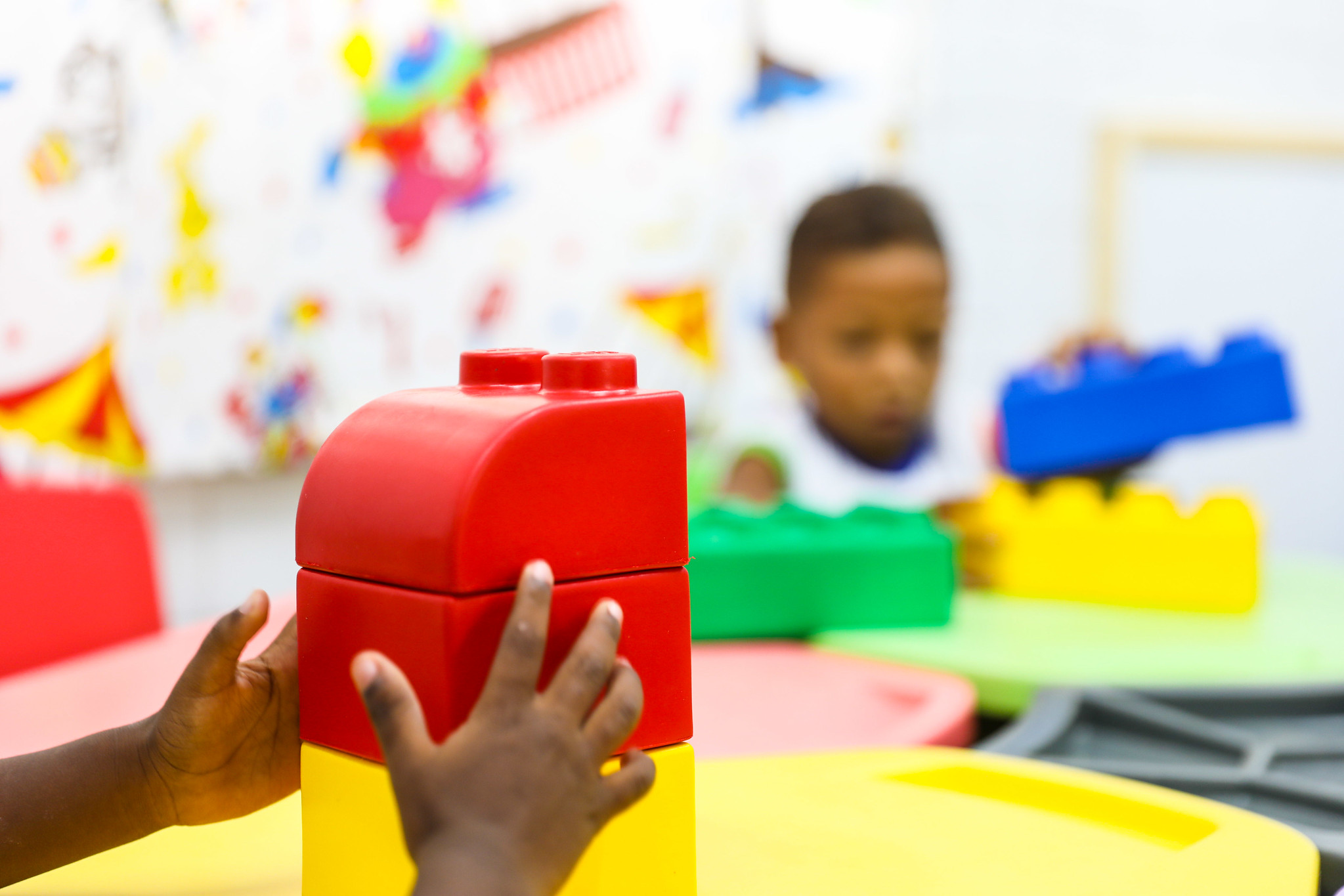 A foto mostra as mãos de uma criança brincando com um grande bloco vermelho de construção. O bloco parece fazer parte de um conjunto de brinquedos semelhantes. O cenário sugere uma área de brincadeira interna ou uma sala de aula, conforme indicado pela decoração colorida e os móveis ao fundo. O foco está na interação da criança com o brinquedo