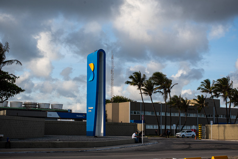 A imagem mostra a fachada da Braskem, em Maceió, com um grande pilar azul com um logotipo amarelo e branco, localizado na frente de um prédio baixo. O céu está parcialmente nublado e há algumas árvores visíveis. Um carro branco está estacionado próximo ao pilar, e uma pessoa pode ser vista sentada perto dele.