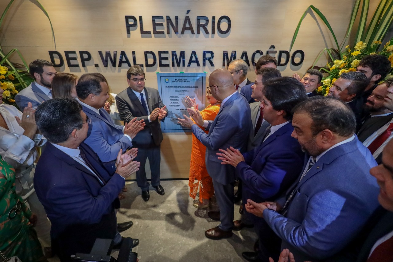 Esta foto mostra um grupo de pessoas em um ambiente interno, em um espaço oficial na Assembleia Legislativa do Piauí. No centro da imagem, há uma mulher vestindo um vestido laranja ao lado do governador Rafael Fonteles, homem branco, de cabelos curtos escuros e óculos, vestindo paletó cinza, camisa clara e gravata azul escuro. As outras pessoas ao redor estão aplaudindo. Ao fundo, há uma placa que diz “PLENÁRIO DEP. WALDEMAR MACÊDO”. Além disso, há flores amarelas decorando o local próximo à placa.
