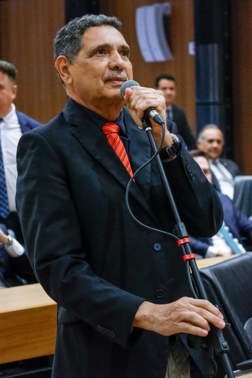 Foto de João Paulo no plenário da Assembleia Legislativa. Ele é um homem negro, idoso, de cabelos curtos e bigode grisalho, falando em pé ao microfone, vestindo paletó e camisa pretas, com gravata vermelha.