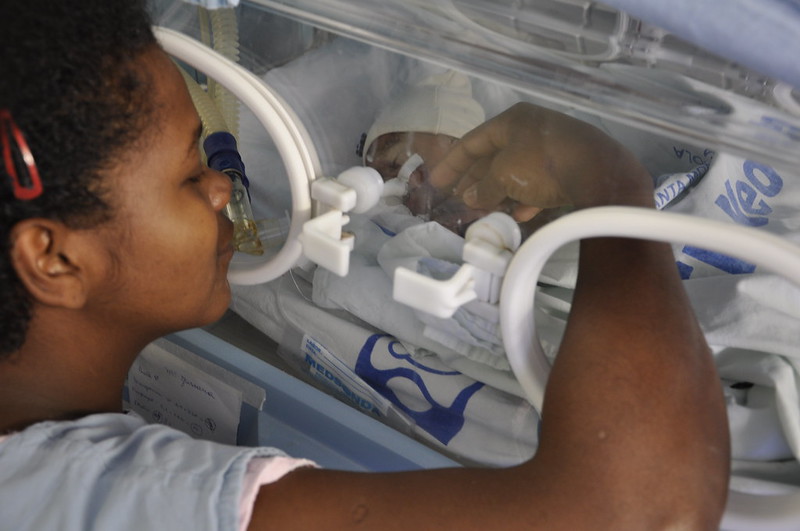 Foto de um bebê negro dentro de um leito de UTI. Ao lado, uma jovem mulher negra, provavelmente a mãe do bebê, toca na criança colocando seu braço por uma abertura do leito.