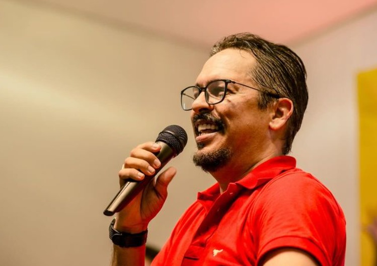 Foto de perfil do Delegado Rossine falando em um microfone, Ele é um homem pardo, de cabelos curtos, de bigode, óculos de aro preto e usando camisa pólo vermelha.