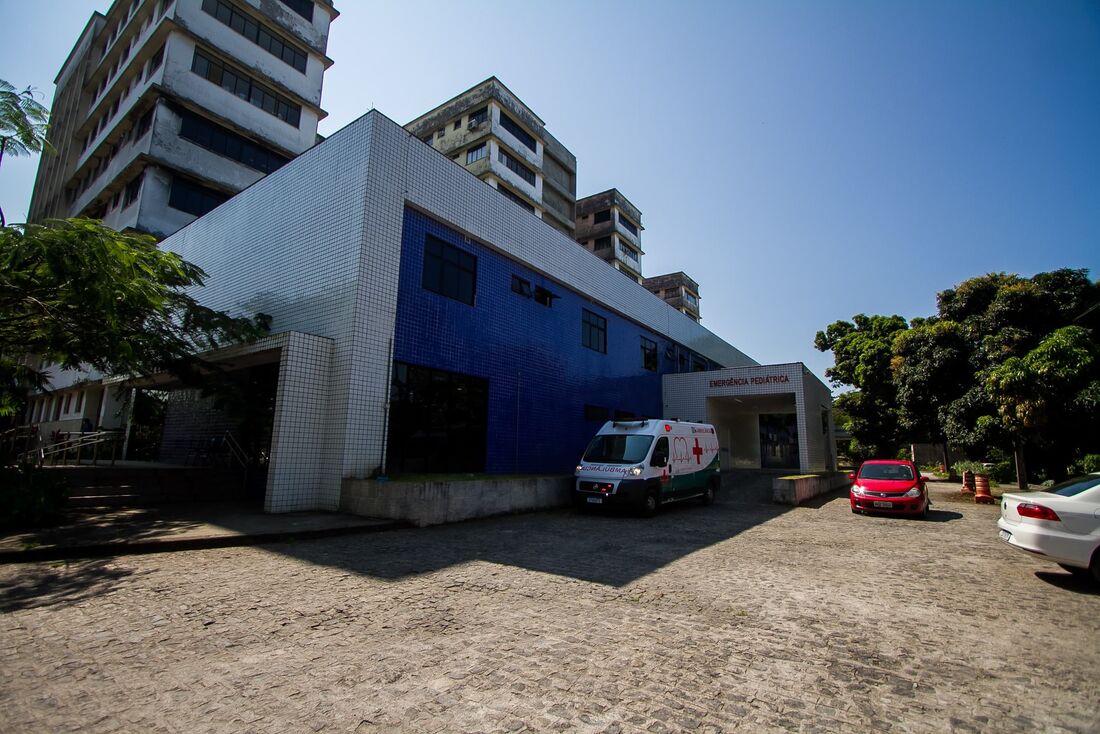 Fachada da emergência pediátrica do hospital Barão de Lucena, em Recife. A imagem mostra um edifício moderno com uma fachada azul no térreo e níveis superiores brancos. Há uma ambulância estacionada em frente ao prédio, sugerindo que possa ser uma instalação médica. Um carro vermelho e um carro branco também estão estacionados nas proximidades. O céu está claro, e há árvores visíveis ao redor da área, indicando um ambiente urbano com alguma vegetação.