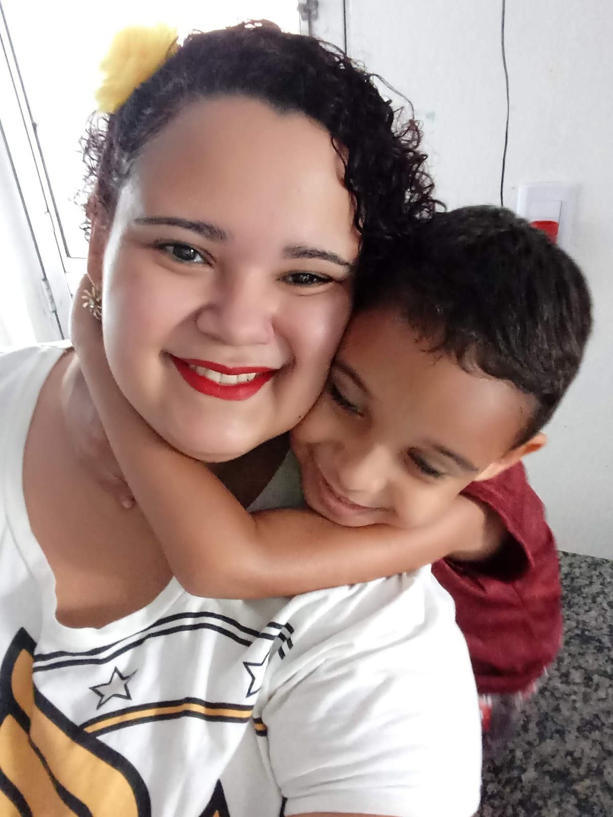 Foto de Cláudia Barrozo e o filho Theo Filipe. Ela é uma mulher parda, de pele clara e cabelos encaracolados, usando camiseta branca. O garoto é negro, de cabelos lisos pretos, que está abraçando a sua mãe com as mãos em torno do pescoço dela.