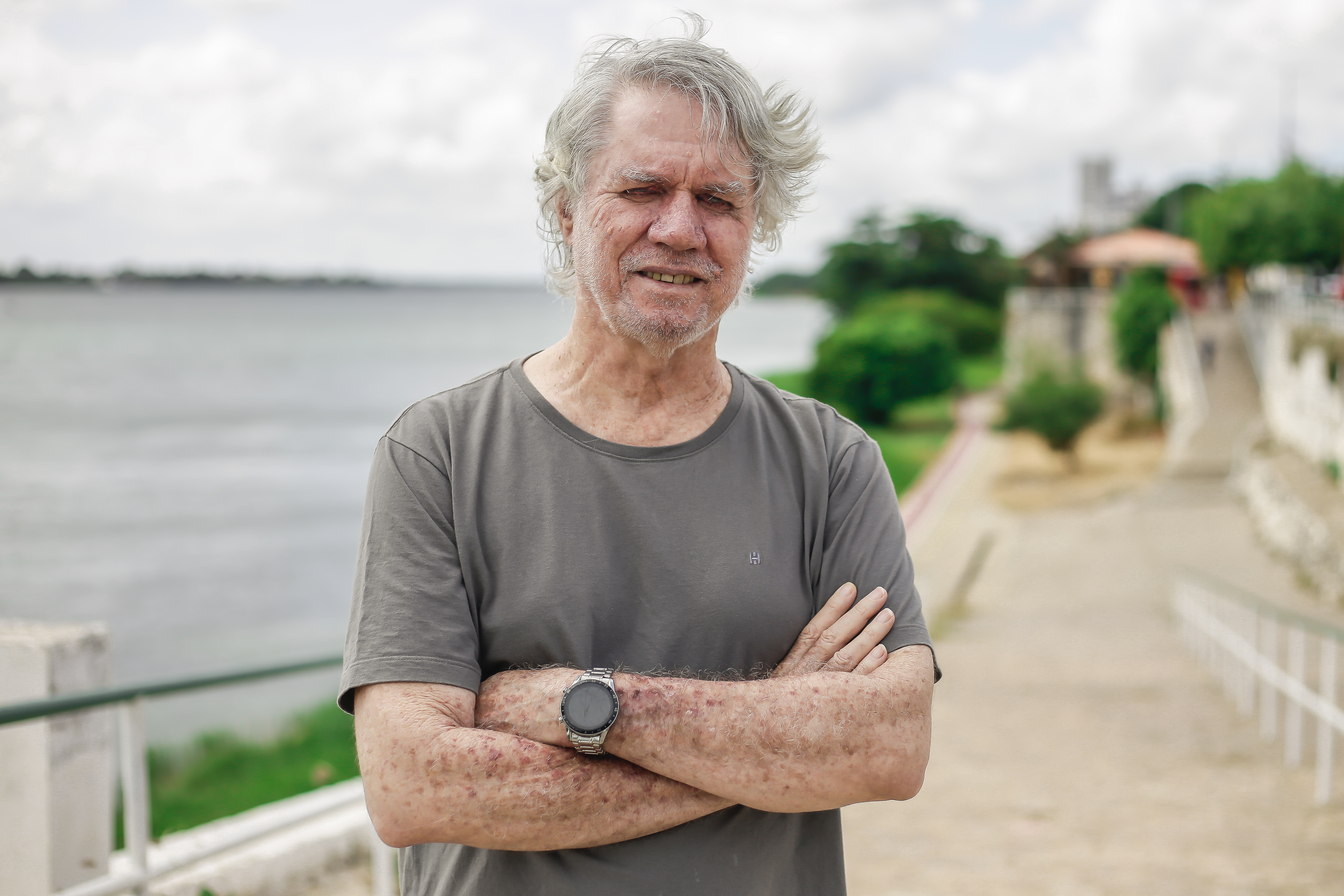 Foto de Roberto Malvezzi, um homem branco, idoso, de fartos cabelos brancos, usando camiseta cinza, de braços cruzados. Ele está em pé numa calçada à beira de um rio, com céu nublado ao fundo.