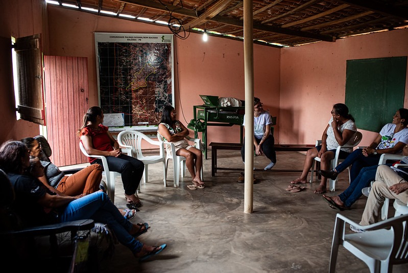 A foto retrata uma reunião em ambiente interno. As pessoas estão sentadas em cadeiras dispostas em um semicírculo. Algumas cadeiras são de plástico e outras de metal. Todos estão voltados para o centro, onde uma pessoa parece estar fazendo uma apresentação ou discurso. O ambiente tem um aspecto rústico, com piso de concreto aparente. Na parede ao fundo, há um grande mapa pendurado. Duas portas são visíveis: uma está aberta, revelando outra sala ou área externa, e a outra está fechada. Um quadro verde de giz também está pendurado em uma das paredes, próximo a uma máquina separadora de sementes.