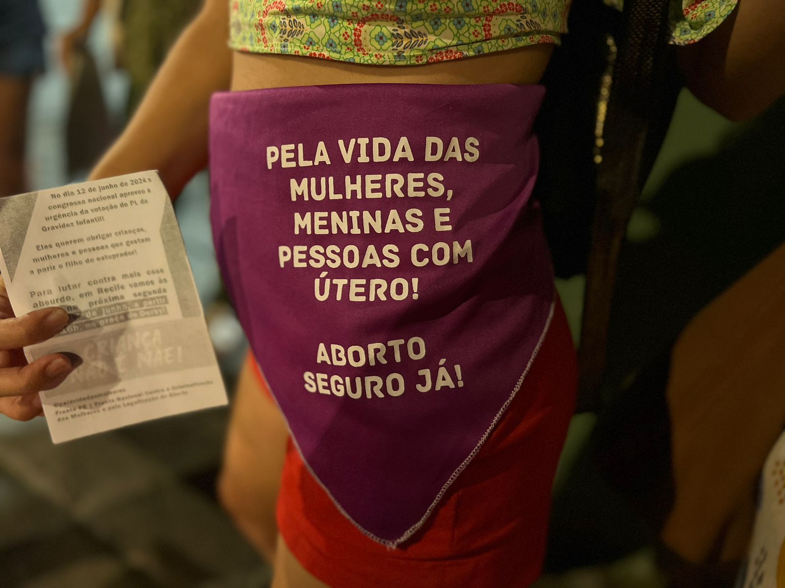 Essa imagem mostra uma pessoa segurando um panfleto em uma das mãos e usando uma bolsa de tecido roxa com texto na frente. O texto na bolsa está em português e se traduz para “Pela vida das mulheres, meninas e pessoas com útero! Aborto seguro agora!” O panfleto parece conter texto, mas está parcialmente obscurecido pela mão da pessoa, tornando-o ilegível.