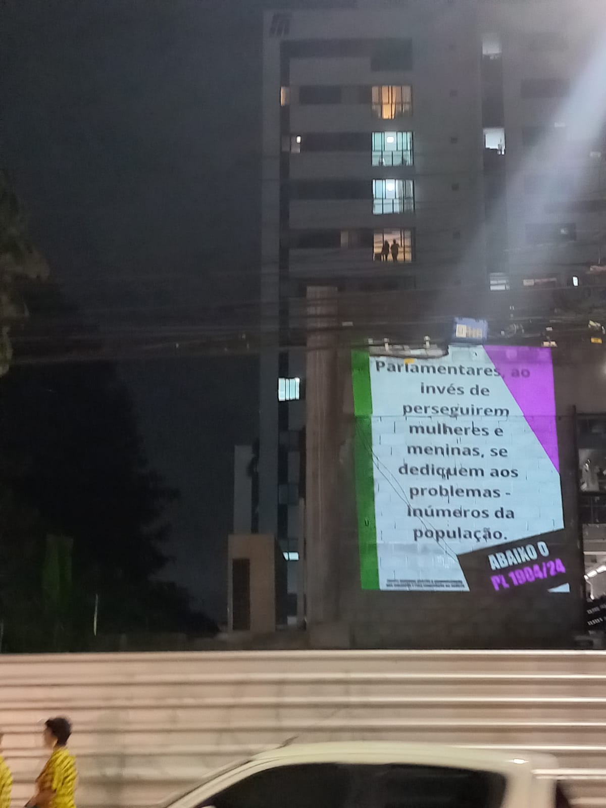 A foto mostra uma cena urbana à noite, com um grande painel luminoso preso à lateral de um prédio de vários andares. O painel contém texto em português, que parece ser uma declaração política ou social. Abaixo do painel, há uma visão parcial de uma rua com veículos em movimento, indicados pelas trilhas de luz devido à exposição prolongada. No lado esquerdo da imagem, há uma pessoa vestindo roupas amarelas, no entanto, apenas parte dessa pessoa é visível devido ao movimento e à posição no quadro. O céu está escuro, sugerindo que é noite. O texto no painel diz: “Parlamentares, ao invés de perseguirem mulheres e meninas dediquem aos problemas da população.”