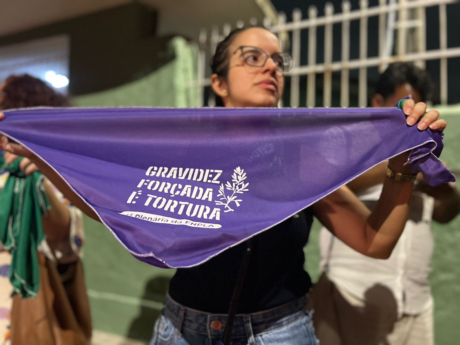 A imagem mostra uma mulher jovem egurando uma faixa roxa com texto em branco. O texto na faixa diz “GRAVIDEZ FORÇADA É TORTURA”, seguido por um texto menor que parece ser uma hashtag, mas não está completamente legível. A jovem está ao ar livre, e há elementos de um ambiente urbano ao fundo, como uma cerca e outra pessoa parcialmente visível.