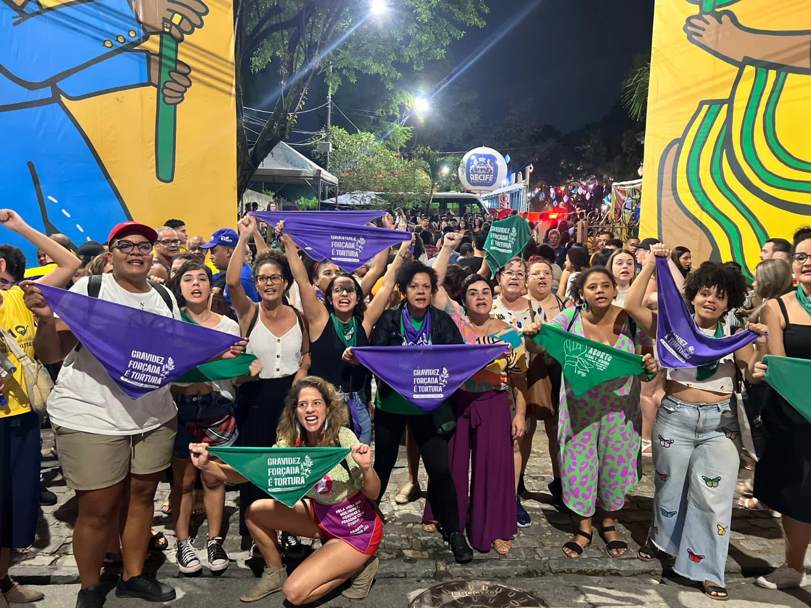 A foto retrata uma cena de rua movimentada durante uma manifestação em defesa do aborto legal. Várias pessoas estão presentes e muitas delas seguram faixas e bandeiras em várias cores, incluindo roxo, verde e amarelo. As faixas contêm textos que defendem os direitos das mulheres e a igualdade. Ao fundo, há um grande mural com cores vibrantes.