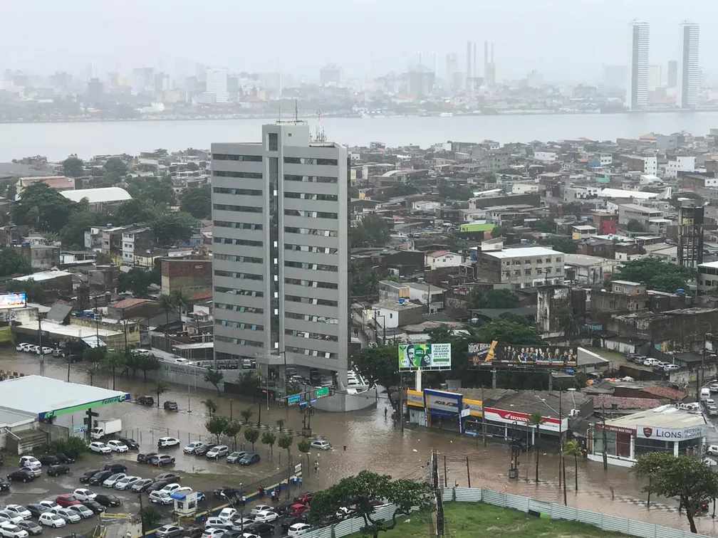 A imagem mostra uma paisagem urbana durante uma enchente significativa. No primeiro plano, há um grande edifício de vários andares com telhado plano, cercado por água que inundou as ruas e submergiu parcialmente os veículos. Além deste edifício, é visível um bairro densamente povoado com várias pequenas construções, também afetadas pelas águas da enchente. Ao fundo, vê-se o horizonte da cidade com prédios mais altos envoltos em névoa, indicando chuva ou neblina. O céu está nublado, e a atmosfera geral parece sombria e úmida.