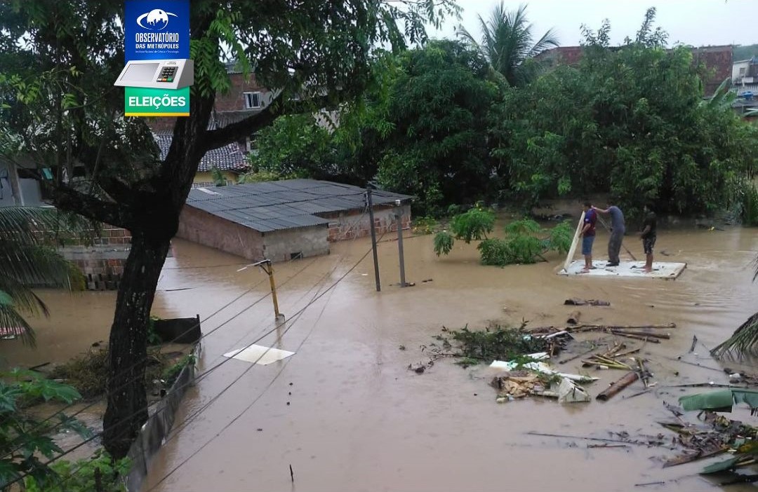 A imagem mostra uma área urbana alagada com água barrenta cobrindo o chão. Duas pessoas estão em uma jangada improvisada feita de tábuas e destroços, aparentemente tentando se deslocar pela água. Várias construções, possivelmente casas ou comércios, estão parcialmente submersas. Há uma placa de rua visível, também parcialmente submersa, indicando que normalmente é uma via pública. No canto superior esquerdo, há uma placa azul com texto branco que diz “Eleições” e Observatório das Metrópoles