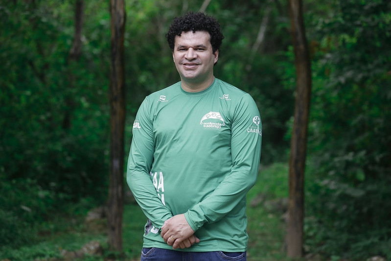 Foto de Gilson Miranda. Ele é um homem jovem, pardo, de cabelos pretos encaracolados, nariz largo, que sorri discretamente para a câmera. Ele veste uma camisa verde de mangas longas do tipo anti UV e tem as mãos juntas na altura do abdômen. Gilson foi fotografado da cintura para cima em um ambiente de mata fechada.