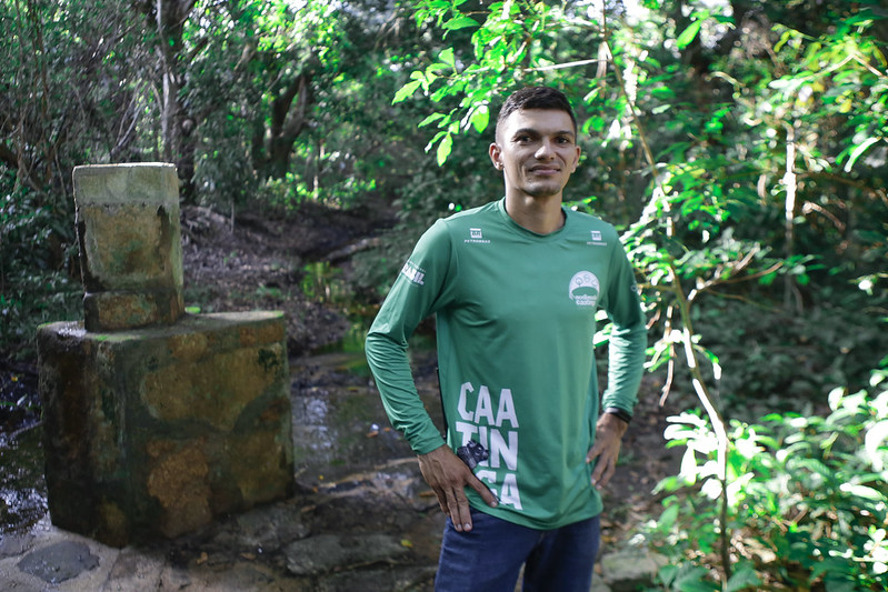 Na foto, vemos Benedito Pereira em pé ao ar livre, em um ambiente natural, possivelmente uma floresta ou parque. Ele é um jovem negro, de cabelos curtos escuros, usando uma camiseta verde de mangas compridas com logotipos e texto em branco. Ele foi fotografado das coxas para cima, olhando fixamente para a câmera, com um semblante sereno. À esquerda da pessoa, há uma estrutura de concreto visível, possivelmente parte de uma fonte de água ou poço, com musgo e descoloração aparentes, sugerindo que está ali há algum tempo. O fundo é preenchido com vegetação exuberante de várias plantas e árvores.