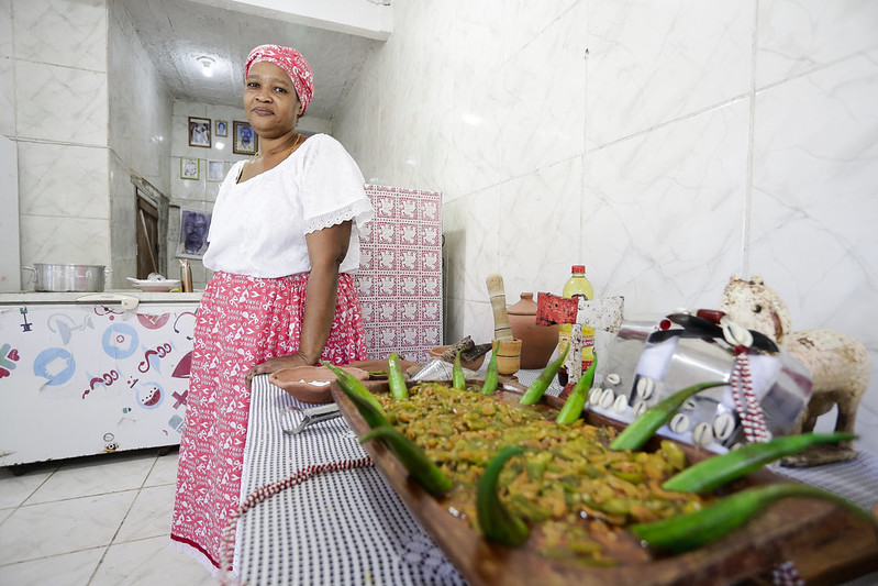 A foto retrata uma cozinha, com paredes revestidas de azulejos brancos. Na frente, há uma grande bandeja de madeira repleta de quiabos verdes, possivelmente um prato tradicional. Ao redor da bandeja, encontram-se vários ingredientes e utensílios de cozinha, incluindo o que parece ser molhos em garrafas, um pilão e almofadas de tecido que podem ser usadas para cobrir alimentos ou como decoração. Ao fundo, está uma mulher olhando para a câmera, usando um adorno na cabeça e uma blusa branca combinada com uma saia vermelha com padrões brancos. Ela está envolvida em tarefas de preparação de alimentos ou cozinha.