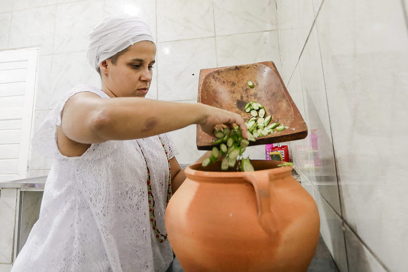 A imagem retrata uma mulher com o semblante sério em pé em uma área que parece ser uma cozinha. Ela está vestindo uma peça de roupa branca que cobre o corpo e a cabeça, lembrando trajes tradicionais. À frente da pessoa, há um grande pote de barro no chão, e ela está segurando uma bandeja marrom inclinada em direção ao pote. Na bandeja, há vegetais verdes cortados que parecem ser quiabo, sendo despejados no pote. O fundo mostra azulejos brancos na parede e armários de cozinha.