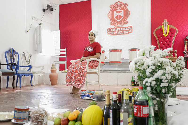 A foto retrata um ambiente interno com foco em uma disposição cerimonial ou ritualística. No centro, há uma mulher vestindo uma camisa vermelha e calças brancas, sentada em um tamborete baixo. Ela é uma mulher negra e jovem, usando um turbante branco na cabeça. Na parede atrás da pessoa, além de tambores, há um grande emblema com um escudo contendo uma cruz, duas machadinhas e texto que parece estar em português. A sala tem paredes vermelhas com padrões decorativos em branco e várias cadeiras ao redor. À frente da pessoa sentada, há uma variedade de objetos dispostos no chão: vários tambores, garrafas de líquidos diversos (incluindo refrigerantes e bebidas alcoólicas), potes, frutas frescas como abacaxis e bananas, e buquês de flores brancas. À esquerda da imagem, há duas cadeiras semelhantes a tronos com estofamento vermelho.