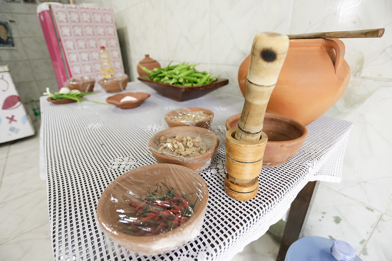 A imagem retrata uma cozinha tradicional com diversos elementos de culinária. À direita da mesa, há uma grande panela de barro com uma colher de pau, e a mesa está coberta por uma toalha de renda branca. Na frente da panela, há um pilão e um almofariz de madeira contendo alguns ingredientes triturados. À esquerda, encontram-se várias tigelas pequenas: uma com pimentas vermelhas e verdes, outra com ervas e uma terceira aparentemente com dentes de alho. Atrás desses itens, há uma variedade de vegetais verdes frescos e o que parece ser uma cesta de plástico rosa ao fundo. A cena sugere preparação para cozinhar com ingredientes frescos usando métodos tradicionais.