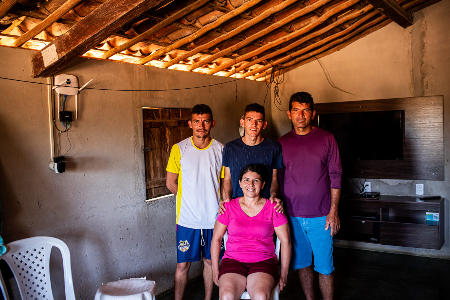 Foto colorida da família de Nelita Pereira Passos, que foi fotograda sentada em uma cadeira de plástico branco. Seu marido e os dois filhos estão em pé por trás dela. O marido é um homem moreno, de pele bronzeada, usando uma camisa roxa de mangas compridas (provavelmente uma antiUV) e bermuda jeans. O filho mais novo é um adolescente magro, de pele clara bronzeada e cabelos escuros e curtos, usando camiseta azul escura. Ele está com as mãos sobre os ombros de Nelita. O filho que aparenta ser mais velho está à esquerda, usando camisa branca com mangas e gola amarelas, com um calção azul. Ele também tem cabelos curtos, mas tem bigode e um cavanhaque ralo. A família foi fotograda dentro de casa, com uma a televisão instalada num painel de madeira escura ao fundo, uma janela fechada na parede à esquerda, modem instalado e na parede e telhado com telhas e caibros de madeira à mostra.