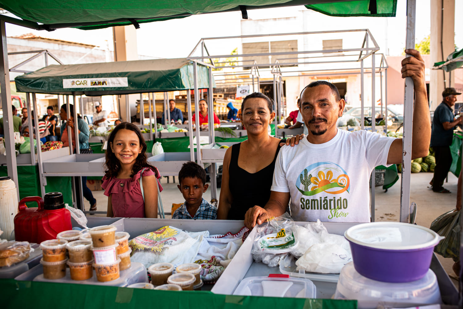 A foto retrata uma cena de mercado com várias pessoas e diversos itens à venda. Na frente, há uma homem negro, maduro, vestindo uma camiseta branca com o texto “Semiarido” e uma ilustração de um cacto e um sol. Ele está com o braço esquerdo esticado, segurando a haste de um barraca de feira. Sua outra mão está apoiada sobre uma mesa repleta de produtos alimentícios em recipientes de plástico. À esquerda, sua esposa, morena, de meia-idade, cabelos presos por trás da cabeça, e usando uma blusa preta. Há duas crianças, um menino negro pequeno e uma menina morena de cabelos lisos e blusa vermelha, estão à esquerda. Ao fundo, mais barracas de mercado podem ser vistas, e outra pessoa vestida de verde está parcialmente visível.