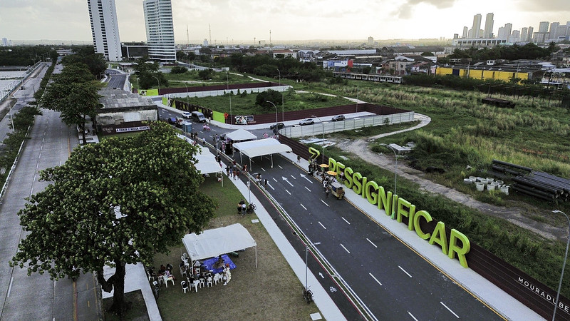 A imagem mostra uma vista aérea de um evento ao ar livre durante o dia. No chão, há um grande texto em negrito que diz “RESSIGNIFICAR”. Abaixo desse texto, há várias tendas brancas alinhadas, com grupos de pessoas ao redor delas. À esquerda das tendas, há uma estrada sem veículos visíveis, e além dessa estrada, há uma área gramada com pouca vegetação. Ao fundo, há prédios altos contra um céu nublado. O cenário parece ser urbano e espaçoso.