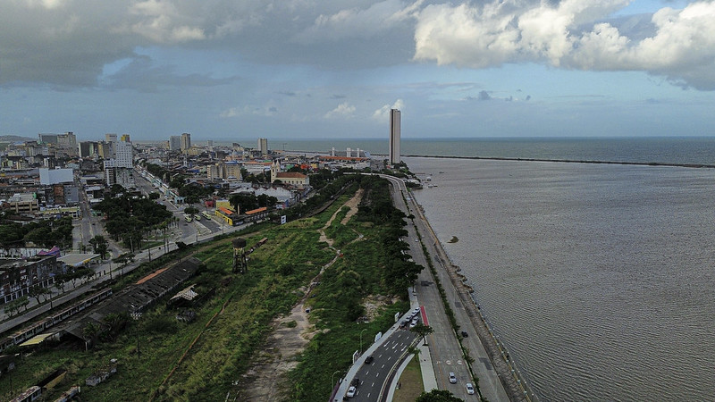 A imagem mostra uma vista aérea do litoral do Recife, na altura do Cais José Estelita. Há uma longa avenida reta que corre paralela à costa, que está à direita da imagem. A costa apresenta um grande corpo de água que se encontra com o horizonte à distância. À esquerda da estrada, há vários edifícios com arquiteturas e alturas variadas, incluindo um arranha-céu particularmente alto e esbelto que se destaca. Entre a estrada e esses edifícios, há uma faixa de espaço verde com árvores e grama. O céu acima mostra nuvens dispersas, sugerindo que pode ser fim de tarde ou início da manhã devido à iluminação suave.