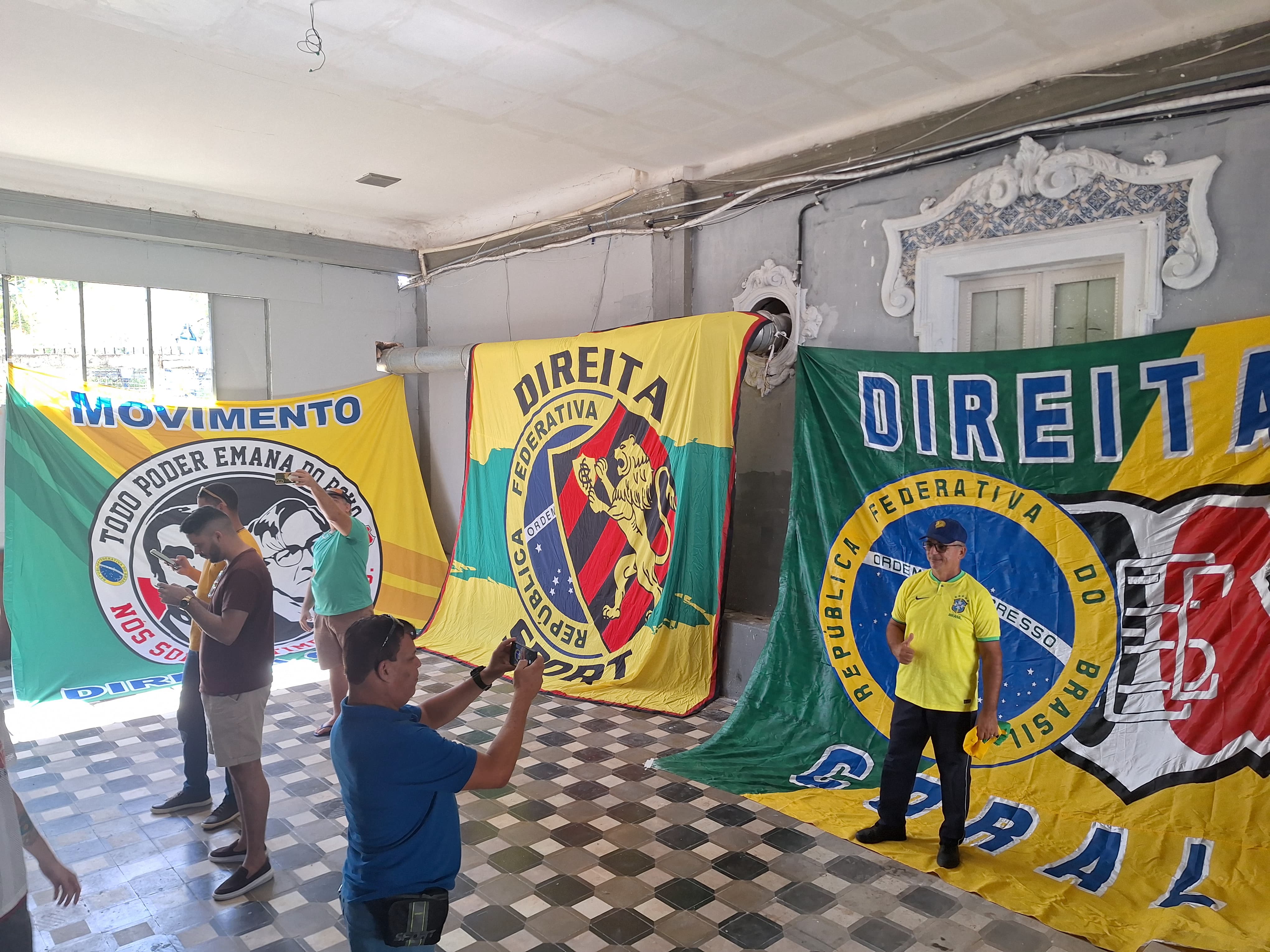 A cena retratada é em um ambiente interno com piso quadriculado e paredes adornadas com molduras decorativas. Três grandes bandeiras estão expostos na sala, cada um com diferentes designs e cores com escudos do Sport Club do Recife e Santa Cruz Futebol Clube. Cinco pessoas estão presentes: uma está em pé à esquerda, segurando um smartphone, aparentemente tirando uma foto ou vídeo dos bandeiras, enquanto a outra está à direita, de costas para a câmera, gesticulando em direção a um dos banners. Os banners contêm texto e símbolos que sugerem que estão relacionados a movimentos políticos da direita.