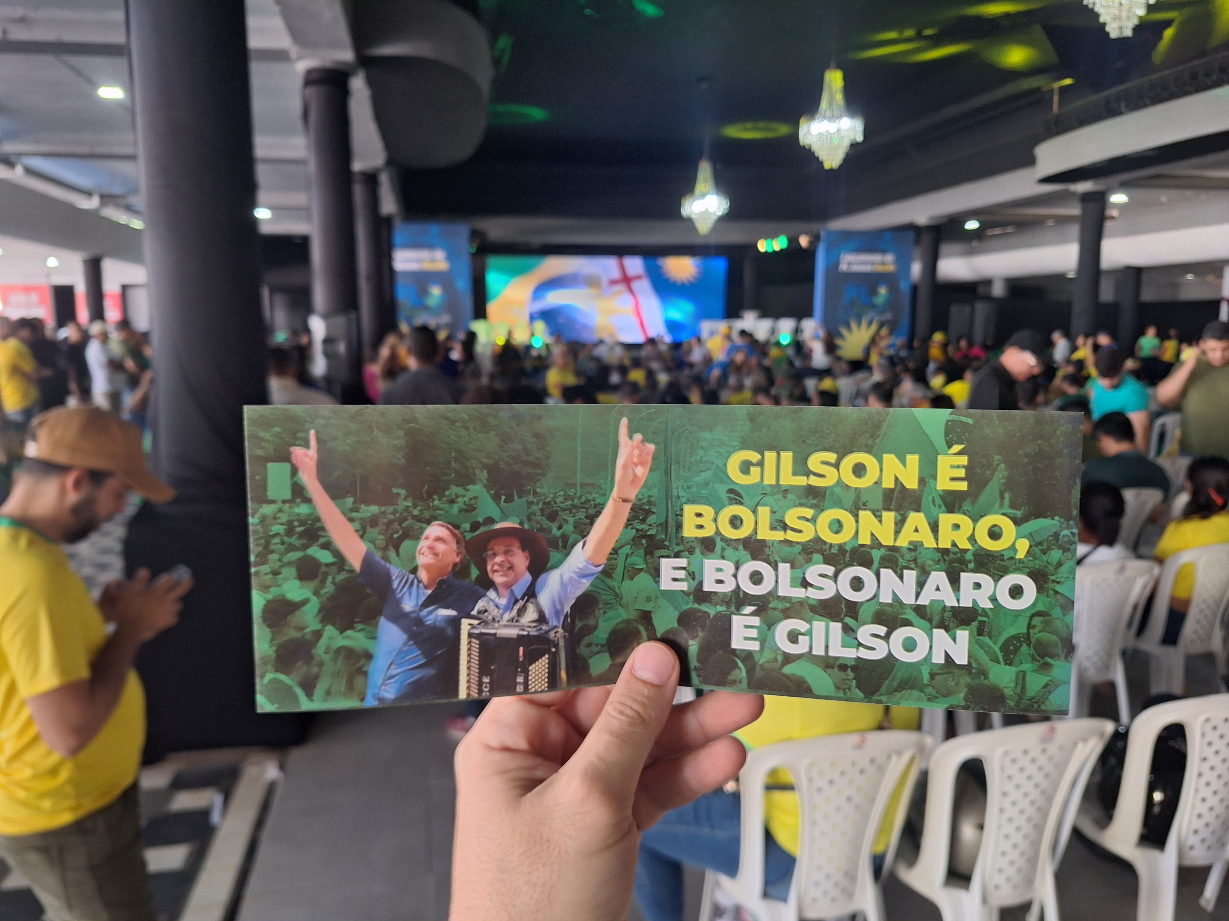 A foto mostra uma mão segurando um adesivo retangular com texto e uma imagem. O adesivo está em foco, enquanto o fundo está levemente desfocado, mas ainda reconhecível. O texto no cartão diz “GILSON É BOLSONARO, E BOLSONARO É GILSON” em letras brancas e em negrito sobre um fundo verde, com pessoas levantando os braços em celebração ou apoio. Ao fundo, parece haver um evento indoor com muitas pessoas vestindo camisas amarelas, sugerindo um evento político ou de campanha. Banners nas cores da bandeira brasileira – verde, amarelo e azul – estão pendurados no teto.