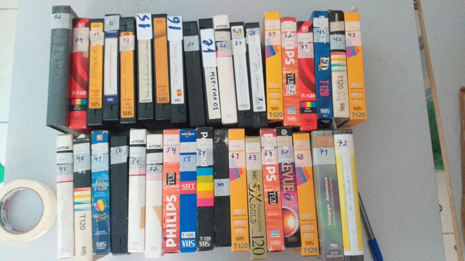 Foto de dezenas de antigas fitas VHS de capas coloridas e numeradas. As fitas estão enfileiradas sobre uma mesa branca, com uma caneta bic azul no canto inferior direito da foto.