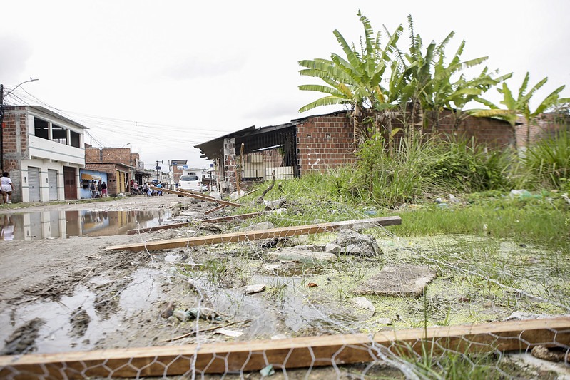 A foto retrata uma cena ao ar livre de uma área inundada com água barrenta cobrindo o chão. Vários prédios estão dos dois lados da enchente, construídos com tijolos e outros materiais, indicando uma área residencial ou comercial. Uma tábua de madeira está colocada sobre o terreno enlameado, possivelmente servindo como uma passarela improvisada. No primeiro plano, uma cerca de metal com arame farpado está jogada no chão. Vegetação, incluindo capim alto e bananeiras, é visível no primeiro plano e no fundo, sugerindo que esta área pode ser propensa a inundações ou estar próxima de uma fonte de água. O céu está nublado, o que poderia implicar chuvas recentes ou iminentes.