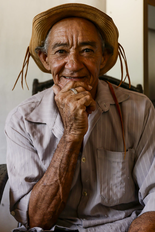 Foto de Nestor Costa, homem idoso, branco de pele bronzeada pelo sol, olhando diretamente para a câmera com a mão direita segurando o queixo. Ele está usando um chapéu de couro de vaqueiro, com fitas nas laterais, vestindo uma camisa cinza claro.