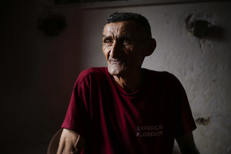 Foto de Alcides Peixinho: ele é um homem negro, de pele curtida pelo sol, cabelos crespos curtos, usando camiseta bordô onde se lê expedição florescer na altura do peito esquerdo. Ele foi fotografado em um ambiente escuro, de paredes descascadas e está olhando para a fonte de luz lateral onde ele e o fotógrafo estão.