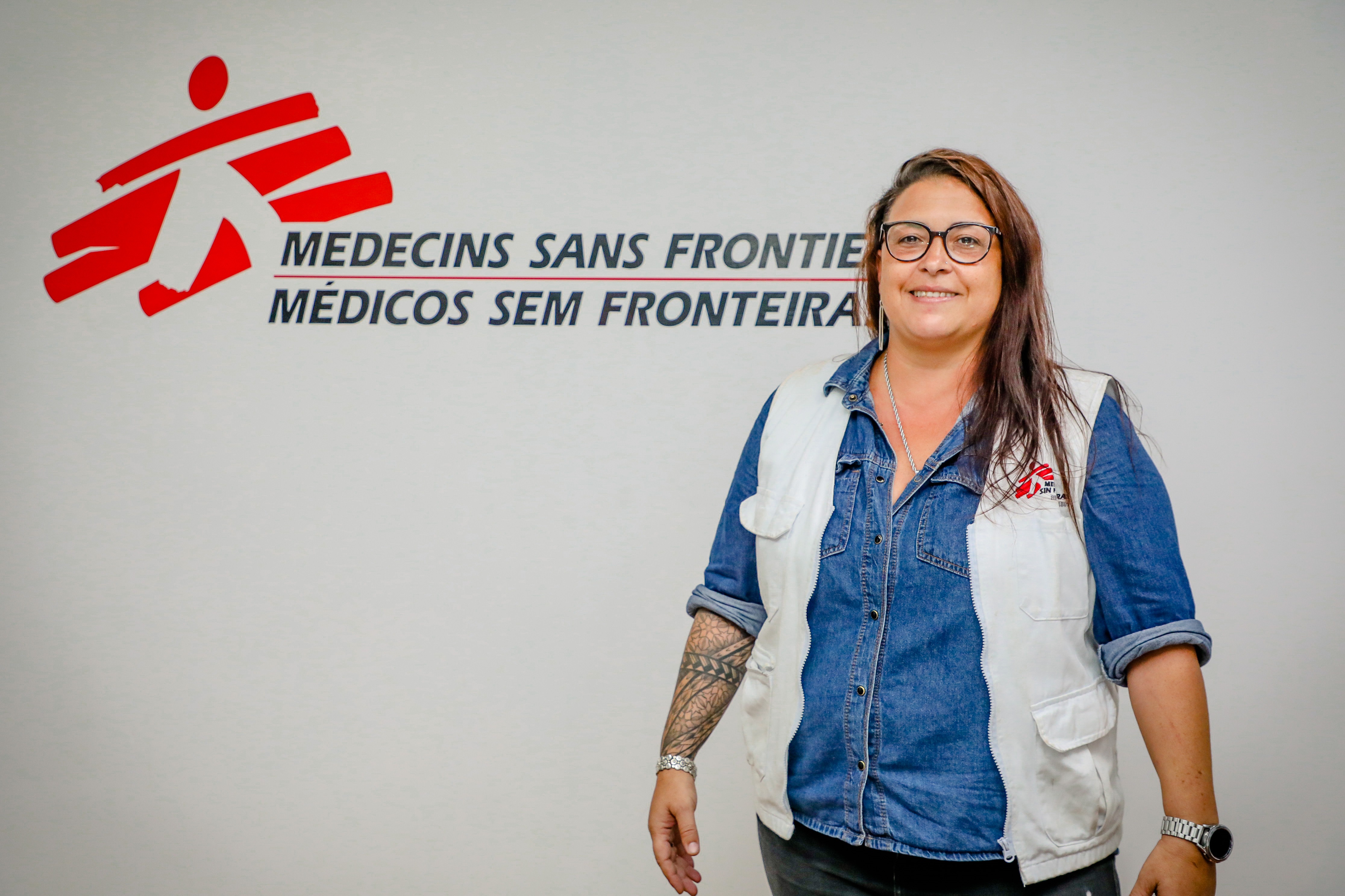 Foto de Lúcia Brum em frente a uma parede com um logotipo e texto. O logotipo consiste em uma figura humana estilizada em vermelho com um recorte branco, seguido por duas linhas de texto. A linha superior diz “MEDECINS SANS FRONTIERES” em letras maiúsculas vermelhas, e a linha inferior diz “MÉDICOS SEM FRONTEIRAS,” em letras maiúsculas azuis. Lúcia Brum está vestindo uma jaqueta jeans azul claro sobre o que parece ser uma camisa branca com um emblema no lado esquerdo, igual ao logotipo na parede, mas menor. Esla é uma mulher branca com cabelo comprido castanho claro que passa dos ombros e tem tatuagens visíveis no braço direito.