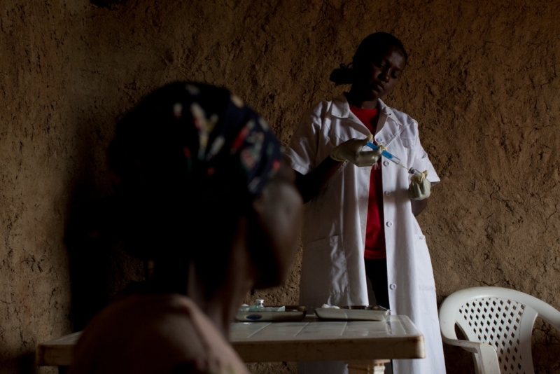 Na foto, paciente com a doença do sono sendo atendida em uma clínica móvel do Médicos Sem Fronteiras em Doromo, na República Democrática do Congo. A imagem mostra um ambiente interno com paredes de barro e iluminação mínima, focando em duas mulheres negras. A mulher em primeiro plano está sentada de costas para a câmera, dificultando a identificação de detalhes específicos. Ao fundo, há outra pessoa vestindo um jaleco branco, sugerindo uma médica. Esta pessoa parece estar manuseando algum tipo de equipamento ou instrumento sobre uma mesa, possivelmente realizando um exame.