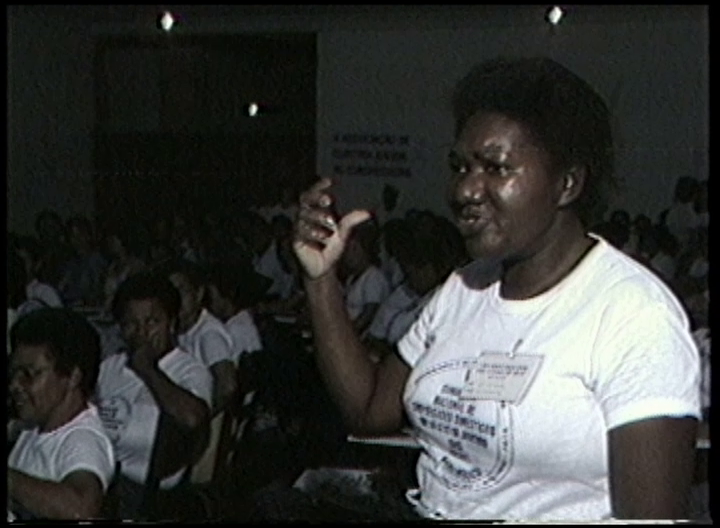 Imagem de baixa resolução de mulher negra de meia idade, gesticulando com a mão direita diante de uma plateia de mulheres sentadas. Ela usa uma camiseta branca com algo escrito e uma crachá preso no tecido da camiseta.