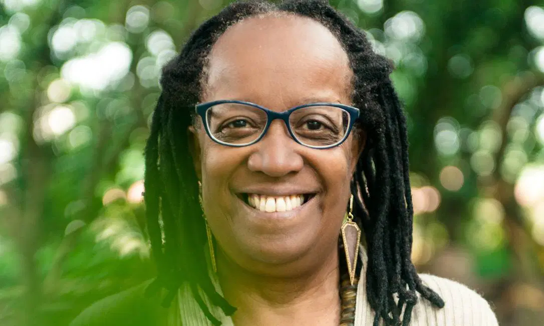 Foto em close de Sueli Carneiro: mulher negra, sorridente, de cabelos trançados e óculos de aro azul, com dois grandes brincos triangulares pendentes.